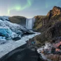 منظر بانورامي ساحر لجبل بركاني نشط في آيسلندا يتدفق منه الحمم البركانية (Lava Flow) نحو نهر جليدي أزرق ضخم (Vatnajökull) يذوب على شاطئ Reynisfjara الأسود الشهير، مع شلالات Gullfoss في الخلفية وفتحات بخار (Geysir) وشفق قطبي (Northern Lights) خافت يظهر في سماء twilight.