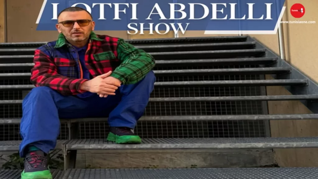 Image of Tunisian comedian Lotfi Abdelli wearing a bold Urban Style outfit, including a multicolor plaid jacket and royal blue trousers, with the text "LOTFI ABDELLI SHOW" at the top. This featured image relates to the latest news about his legal case and his unique fashion choices.